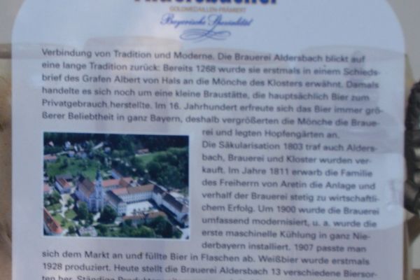 Auf einem Plakat wird die Geschichte der Brauerei Aldersbach beschrieben. In den Text eingebettet ist eine Fotografie der Brauerei mit Klosterkirche.