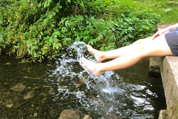 Auf einem steinernen Steg sitzt eine Person in kurzen Hosen und lässt ihre nackten Beine in das Flussbett baumeln. Die Füße spritzen dabei das Wasser empor. Hinter dem grünen Uferrand ist eine Steinmauer.