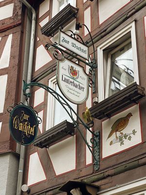 Blick auf ein Detail an einem Fachwerkhaus neben der Eingangstür. Zu sehen sind schmiedeeiserne Metallschilder, deren Gestänge kunstvoll gerabeitet ist. Auf einem rechteckigen Schild steht Zur Wachtel, darunter auf einm wappenförmigen ist ein Brauereilogo zu erkennen mit dem Schriftzug Lauterbacher und auf einem dritten, schwarzen Schild, das rund ist, steht in Gold Zur Wachtel geschrieben. In einem weißen Gefach des braunen Fachwerks ist eine Wachtel rechts neben den Schildern gemalt.