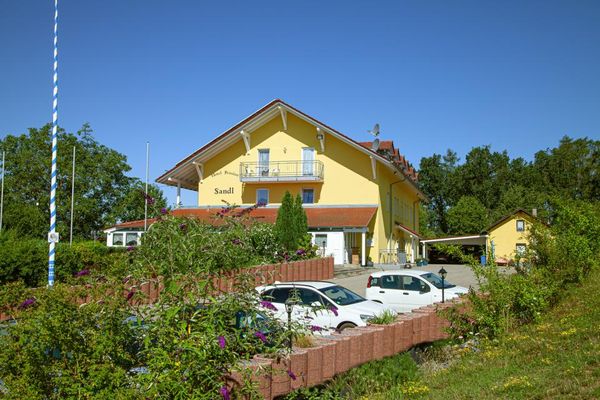 Das gelbe Gebäude mit Dachgauben trägt die Aufschrift "Hotel Pension Sandl" und verfügt über eine große, überdachte und geschlossene Terrasse. Ziegelfarbige Pflanzringe trennen den Parkplatz mit geparkten Autos von einer Wiese mit blühenden Sträuchern. Im Hintergrund stehen ein Nebengebäude und hohe Laubbäume.
