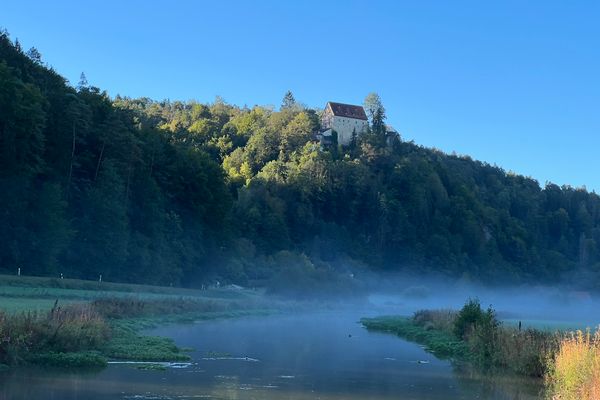 Ein Burg thront erhöt auf einem Hügel. Unten im Tal liegt Nebel auf dem Fluss.