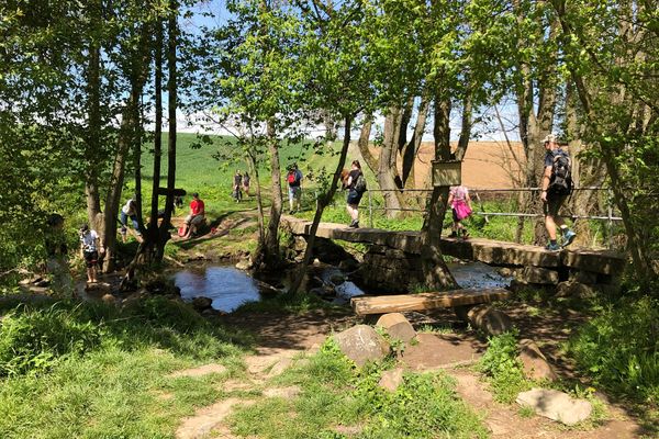 Eine Bachquerung über eine schmale Steinbrücke mit einseitigem Geländer. Vor der Brücke steht eine einfache Bank aus Holzstämmen und Balken. Überall sind Menschen unterwegs, auch im Wasser. Auf einem Schild an einem Baum steht Naturkneippen.