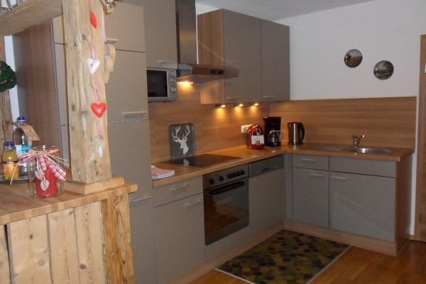 Eine L-förmige, graue Küchenzeile mit Mikrowelle und weiteren Elektrogeräten, daran schließt sich eine Holzkommode mit Überbau an, auf der dekorative Gegenstände stehen.