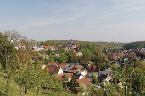 Der Blick geht in eine Talsenke, umgeben von Wiesen, Feldern und Wäldern, in der der Ort eingebettet ist. Linkerhand liegt auf einem Bergsporn ein kleines Schloss.