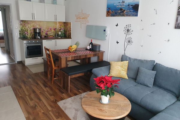 Wohnraum mit kleiner, weißer Küchenzeile, einem Holzesstisch samt zwei Bänken und einem Stuhl. Dahinter ein graues Ecksofa und ein dunkler, runder Tisch mit Holzplatte. An der Wand befinden sich Bilder und verschiedene Dekomalerei.