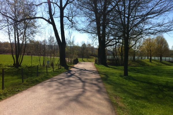 Der Betrachter steht auf einer Landstraße die vor ihm geradeaus einen Bach überbrückt. An der Brücke stehen beidseitig hohe Laubbäume.