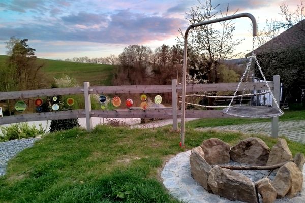 Grillplatz mit Grillrost der über der Feuerstelle an einer Kette befestigt ist. Im Hintergrund sind Wiesen und Bäume zu sehen, sowie ein Holzzaun mit bunter Deko aus Glas