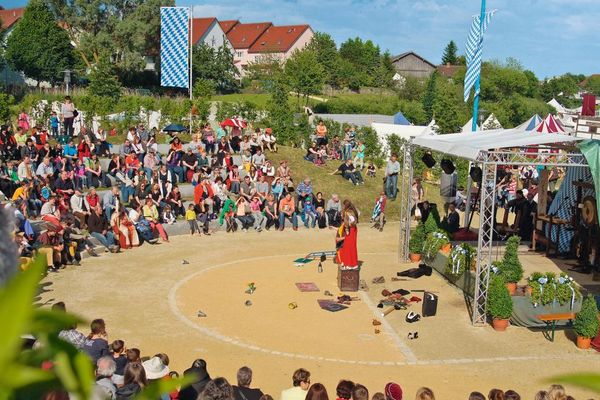 Auf ansteigenden Sitzreihen verfolgen viele Zuschauer ein Schauspiel der Natur-Freilichtbühne. Daneben wehen zwei Bayernfahnen. Im Hintergrund einige Laubbäume und Gebäude.