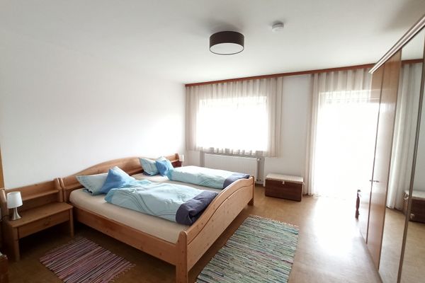 Ein Doppelbett im Holzrahmen mit zwei Nachttischen steht im Raum. Gegenüber befindet sich ein Kleiderschrank mit Spiegeltüren. Vor dem Bett liegen zwei Flickenteppiche. Helle Gardinen hängen an Fenster und Balkontür.