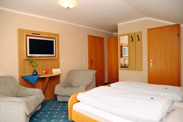 Zimmer mit Doppelbett im Holzrahmen, Sitzecke mit zwei Sesseln, Garderobe und Spiegel. In einer fast deckenhohen Holzwand sind Flachbildfernseher und Tisch eingebaut.
