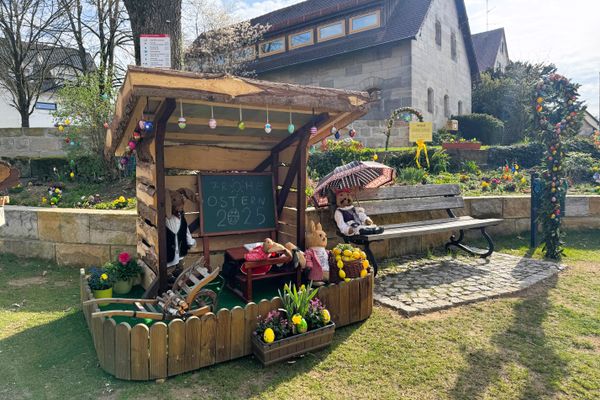 Tafel, Schulbank mit drei Häschen, Hasenlehrer unter kleiner Holzüberdachung. Daneben Bank mit Osterhasen unter einem Regenschirm. Alles geschmückt mit Ostereiern und Frühlingsblumen.