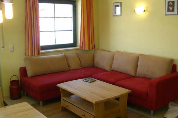 Rotes Ecksofa mit grauen Rückenpolstern, davor ein Holztisch. Am Fenster hängen rotgemusterte Vorhänge. Zwischen zwei Wandbildern leuchtet eine Lampe.