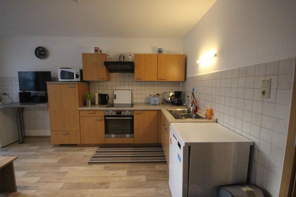 Wohnküche mit L-förmiger Holzküchenzeile, separater Spülmaschine und verschiedenen Elektrogeräten. Neben der Küchenzeile steht ein TV-Gerät auf einer grauen Arbeitsplatte.