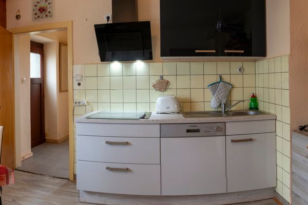 Das Bild zeigt eine Küche. Links befindet sich eine offene Tür zu einem Flur. In der Mitte ist eine weiße Küchenzeile mit Spülbecken, Toaster und Geschirrspüler. Darüber sind gelbe Fliesen und ein schwarzer Dunstabzug. Rechts im Bild ist eine Kaffeemaschine.