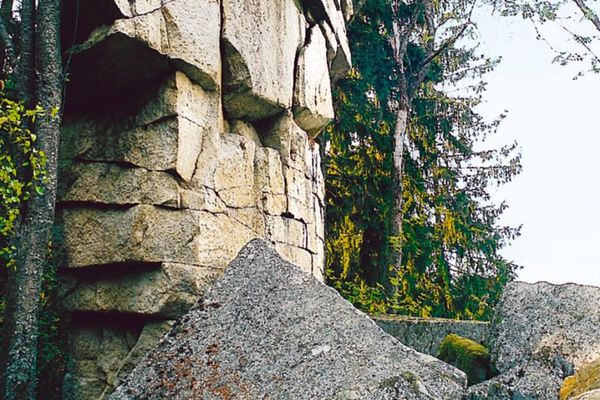 Hinter einigen Felsblöcken und vor Laubbäumen steht eine aus unregelmäßigen Steinen gebaute Mauer.
