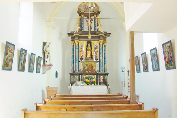 Heller Altarraum mit einreihigen Kirchenbänken. An den Seitenwänden Kreuzwegstationen, links eine Heiligenfigur.