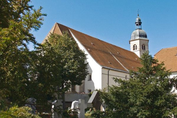 Unter blauem Himmel im Vordergrund Grabsteine unter hohen Bäumen, dahinter ein Teil der Wallfahrtskirche Bogenberg mit Kirchturm.