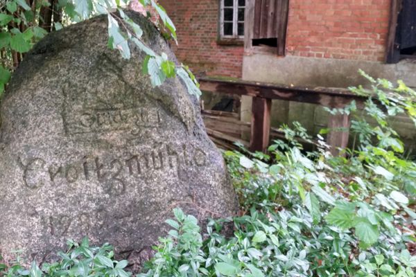 Historischer Gedenkstein an der Kroetzmühle vor einem alten Backsteingebäude, umgeben von Bäumen und Sträuchern.