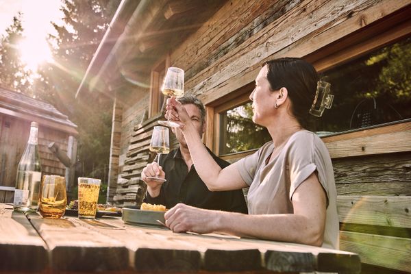Ein Paar mittleren Alters sitzt an einem Holztisch vor einer Holzhütte im Freien, umgeben von Grün. Sie prosten sich mit Weingläsern zu und auf dem Tisch steht Essen vor ihnen.