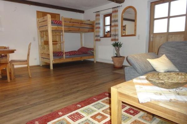 Im Schlafwohnraum mit Sprossenfenster und -tür steht ein kieferfarbenes Stockbett. Essbereich und Wohnteil mit Sofa und Tisch sind ebenfalls sichtbar. Teppich und Vorhang sind bunt gemustert. An der Wand hängt ein holzgerahmter Spiegel und dunkle Sichtbalken zieren die helle Decke.