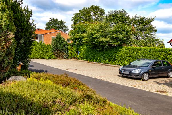 Ein grauer Kleinwagen steht auf einem gepflasterten Parkplatz, der von einer grünen Hecke begrenzt wird. Im Vordergrund verläuft ein asphaltierter Weg an einem Beet mit Heidepflanzen vorbei. Im Hintergrund sind Bäume und die Dächer von rot geklinkerten Wohnhäusern unter einem blauen Himmel mit Wolken zu sehen.