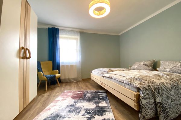 In einem Raum steht ein breites Doppelbett mit einem Holzrahmen. Die Bettwäsche ist hell und mit dunklen, gepunkteten Kreisen verziert. Gegenüber dem Bett befindet sich ein heller Schrank, daneben ein gelber Stuhl. Auf dem Fußboden liegt ein Teppich mit einem rot-schwarzen Muster. Das Fenster ist mit einer weißen Gardine bedeckt, darüber hängt ein bodenlanger, blauer Vorhang.