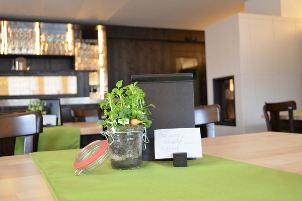 Auf dem Bild ist der Tresen, sowie der Kamin im Hintergrund zu sehen. Im Vordergrund des Bildes befindet sich die Tischdekoration, die auf einem hellen Holztisch steht.