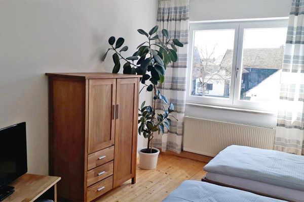 Vor zwei Einzelbetten stehen nebeneinander ein TV-Tisch mit Fernseher, eine Holzkommode und eine Grünpflanze. Bodenlange, helle Vorhänge mit Streifenmuster hängen vor dem Doppelfenster.