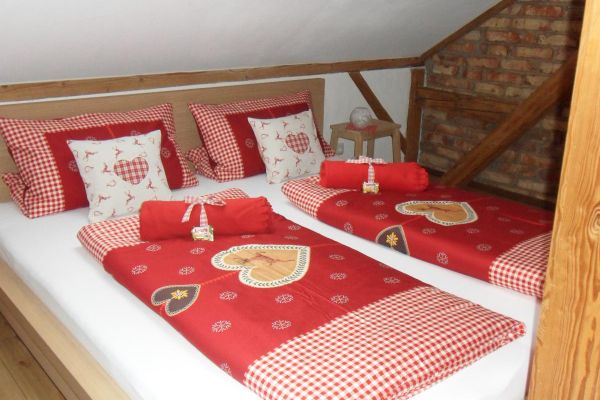 Auf einem Holzfußboden steht unter einer Mansarde mit Dachfenster ein Doppelbett mit Holzrahmen und rotgemusterter Bettwäsche im Landhausstil; daneben eine rustikale Mauersteinwand.