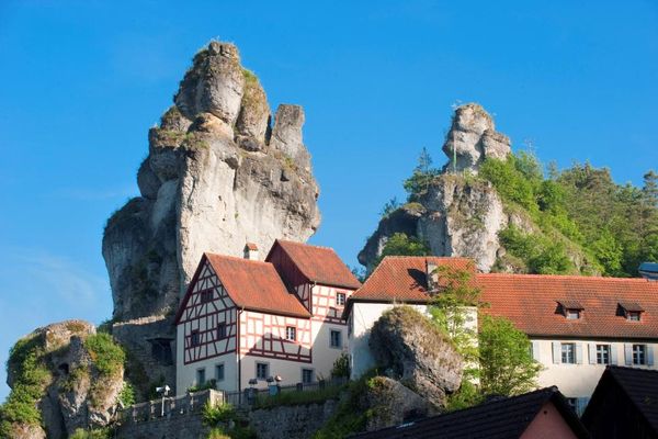 Blick auf ein rot weißes Fachwerkensemble. Im Hintergrund erheben sich zwei mächtige Felstürme.