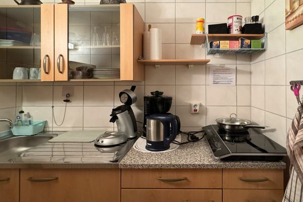 Kleine Küche mit Spüle, Arbeitsfläche, Kaffeemaschine, Wasserkocher und Herdplatte, umgeben von gefliesten Wänden und Hängeschränken.