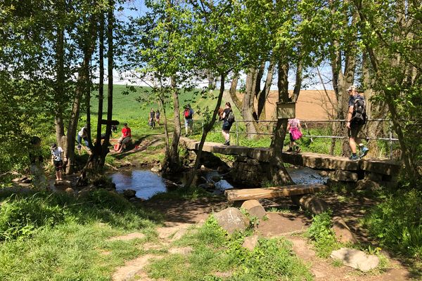 Eine Bachquerung über eine schmale Steinbrücke mit einseitigem Geländer. Vor der Brücke steht eine einfache Bank aus Holzstämmen und Balken. Überall sind Menschen unterwegs, auch im Wasser. Auf einem Schild an einem Baum steht Naturkneippen.