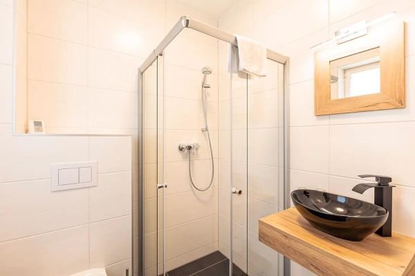 Bad mit Dusche aus Glas, vor der Dusche Waschtisch aus Holz mit schwarzem Waschbecken, darüber ein kleines Fenster, links ist noch ein Teil des WCs zu sehen.