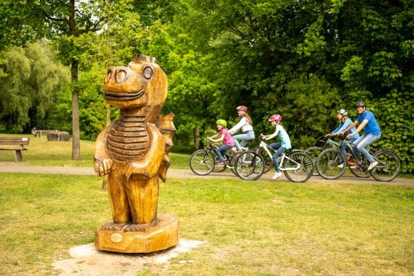 Drachen-Skulptur aus Baumstamm am Drachen-Spielpark, dahinter ankommende Familie mit Rädern