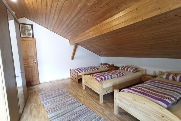 Unter einer holzvertäfelten Dachschräge stehen drei Betten mit Holzrahmen, dazwischen zwei Nachttische. Gegenüber befindet sich ein Kleiderschrank mit mittiger Spiegeltür. Vor den Betten liegt ein Flickenteppichläufer.