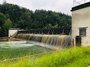 Der Betrachter blickt vom Ufer aus auf die kleine Lechstaustufe über welche das Wasser des Lechs läuft.