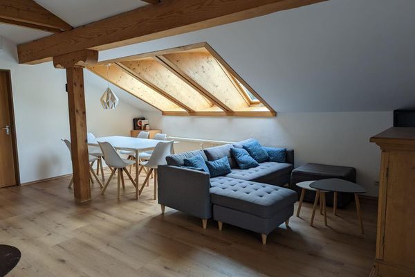 Mansardenwohnraum mit sichtbaren Stütz- und Deckenbalken. Unter drei Dachfenstern steht eine weiße Essgruppe für sechs Personen, daneben ein graues Ecksofa mit Tisch und Hocker vor einer Holzkommode.