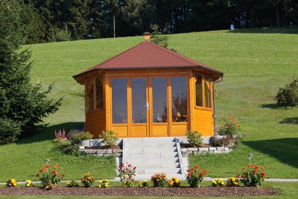 In einer gepflegten Wiese führt eine Steintreppe zu einem Holzpavillon mit vielen Fenstern. Dahinter beginnt der Wald. Vor dem Pavillon blühen bunte Blumen in Beeten, die mit Rindenmulch ausgestreut sind.