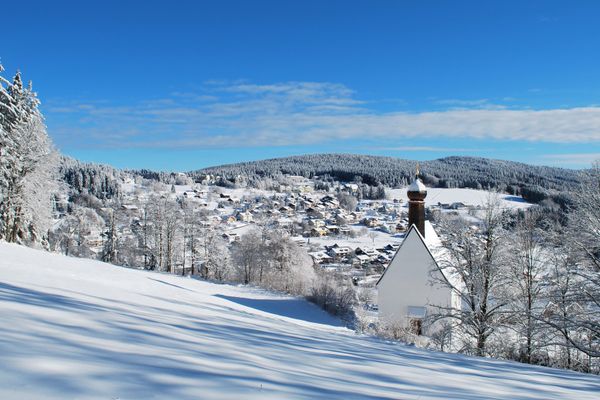 In wunderschöner, verschneiter, sonniger Wald- und Winterlandschaft liegt die Leonhardikapelle und dahinter das Dorf Sankt Englmar, überragt von den schneebedeckten Bäumen der Waldkette.