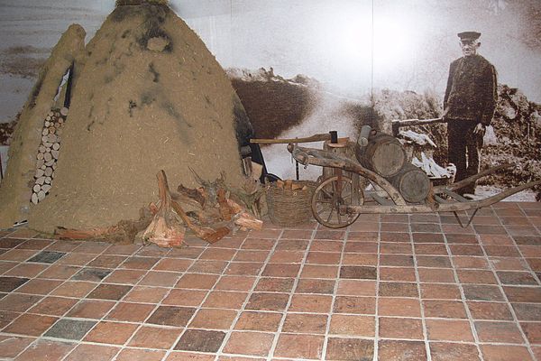 Im Ausstellungsraum ist eine Kohlemeiler aufgebaut, davor steht eine große, hölzerne Schubkarre mit Fässern und gestapeltes Holz. Die Wand dahinter ist ein großes historisches Bild eines Arbeiters, der Holz hackt.