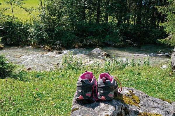 Ein paar rosa-graue Wanderschuhe steht im rechten Vordergrund auf einem Basaltstein, der auf einer Wiese liegt. Quer durch die Mitte des Bildes fließt ein Bach sprudelnd über das steinerne Flussbett. Das gegenüberliegenden Ufer ist dicht mit Gebüsch und Bäumen bewachsen. Eine Wiese blitzt durch das Gestrüpp.
