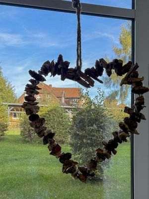 Herzförmige Dekoration aus kleinen Holzstücken und Zapfen hängt vor einem Fenster. Draußen sind Rasen, Sträucher und ein Gebäude zu sehen.