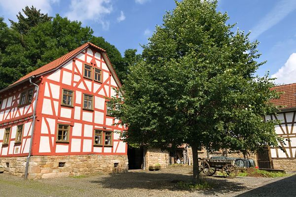 Eine Hofreite mit einem roten Fachwerkhaus links und rechter Hand anschließendem Wirtschaftsgebäude. Dieses hat einen steinernen Sockel, der rechte Flügel des Stalls ist in Fachwerkbauweise errichtet worden. Im geplfasterten Hof davor steht ein hölzerner Leiterwagen unter einem mächtigen Laubbaum, der ganz zentral das Bild einnimmt. Es ist wunderschönes, sommerliches Wetter.