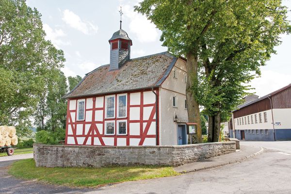 Eine kleine Fachwerkkirche mit roten Balken und einem grau verschieferten Haubendachreiter mit Metallkreuz. Vor dem Eingangsportal stehen zwei große Bäume. Das Umfeld wirkt sehr ländlich, ein Anhänger mit Heuballen steht links im Hintergrun.