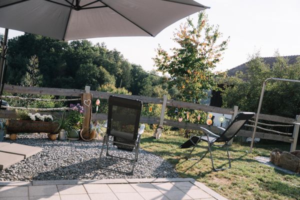 Terrasse mit zwei Liegestühlen und einem Sonnenschirm in grau. Im Hintergrund viele Bäume und Deko sowie eine Katze die am Zaun sitzt.