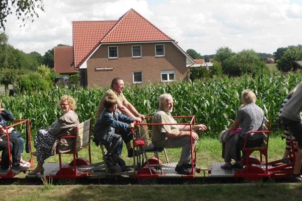 Ein Draisinentriebwagen während der Fahrt auf dem einige Menschen platz gefunden haben. Sie fahren an einem Maisfeld vorbei