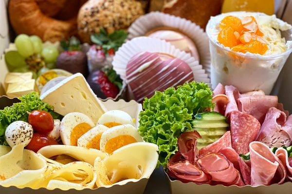 Auf dem Bild ist eine Kiste mit verschiedenen Frühstücksprodukten, wie Gebäck (Hörnchen, Körnersemmeln, Laugengebäck), Schokofrüchte, Desserts, sowie Käse, Wurst und Eier zu sehen.