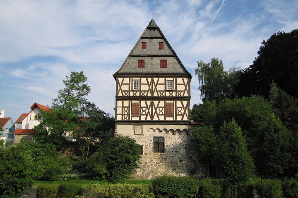 Umwachsene von dichtem Grün steht auf einem massiven steinernen Unterbau ein viergeschossiger Fachwerkaufbau. Der Himmel ist blau, das Wetter gut.
