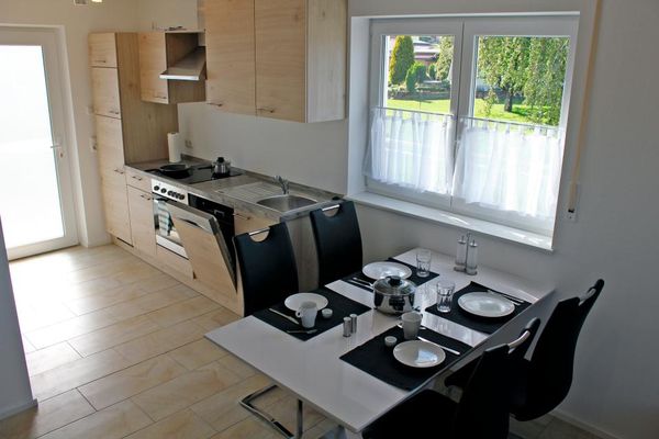 Küchenzeile aus hellem Holz, daneben ein Fenster mit Bistrogardine und davor stehen ein weißer , gedeckter Esstisch und vier schwarze Stühle.