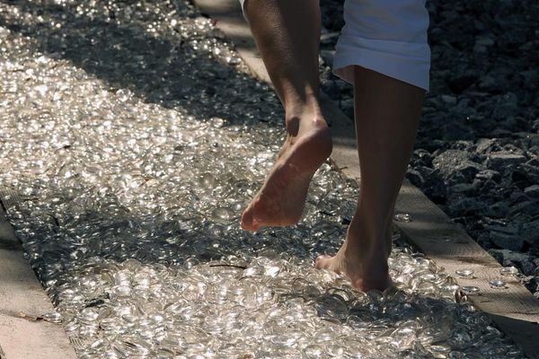 Auf einem durch Holzbalken eingefassten und mit Glasscherben befülltem Weg sieht man nur vom Knie abwärts ein paar Beine, die barfuß auf den Scherben gehen.